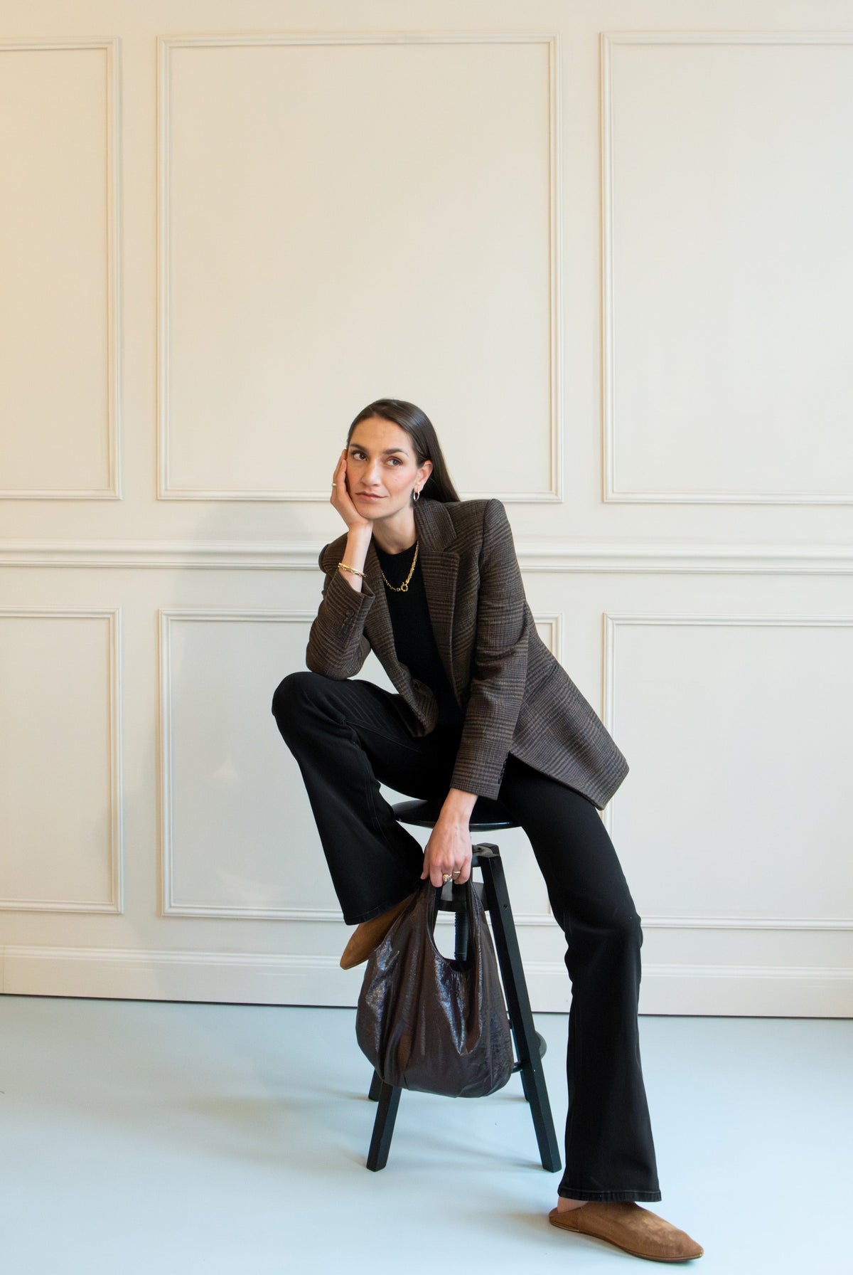 Stylish woman in a checkered blazer and black pants, seated on a stool in an elegant, cream-paneled room.