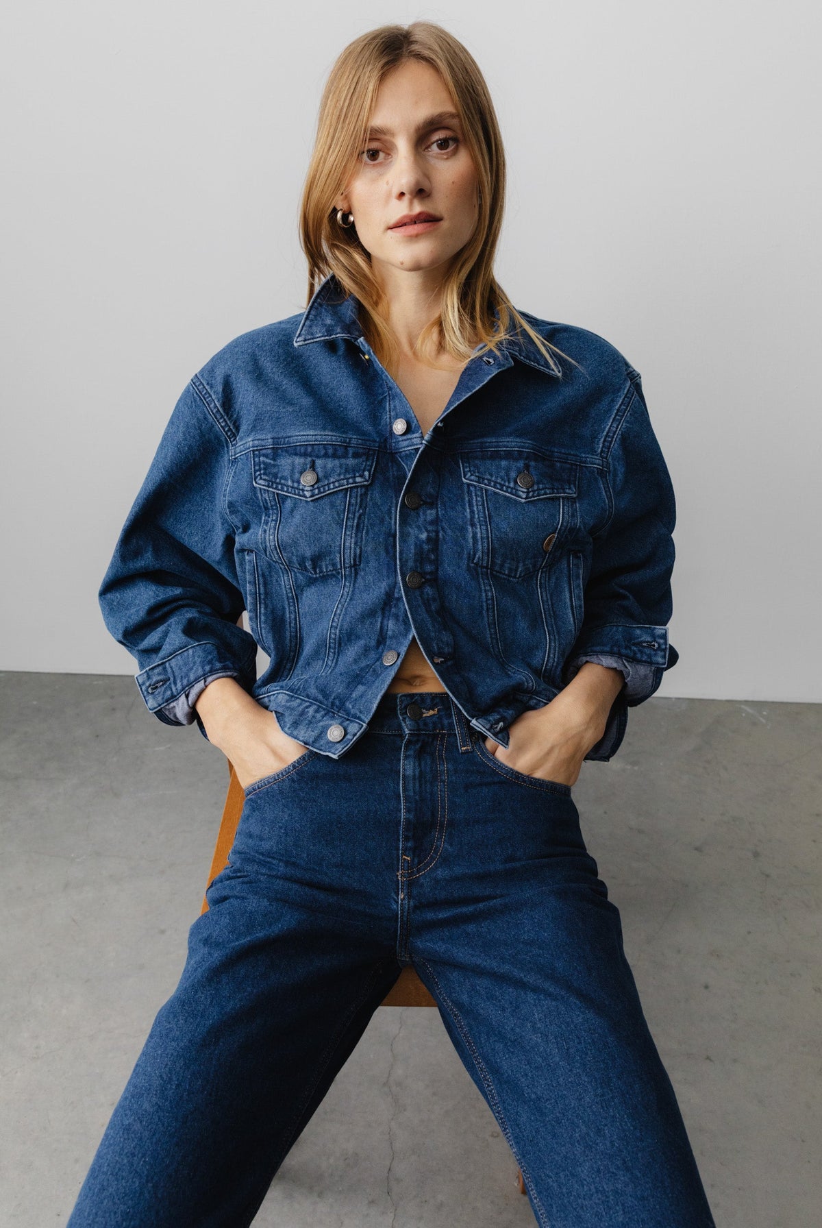 Woman wearing a classic blue denim jacket with matching high-waisted jeans, sitting on a wooden chair against a plain backdrop.