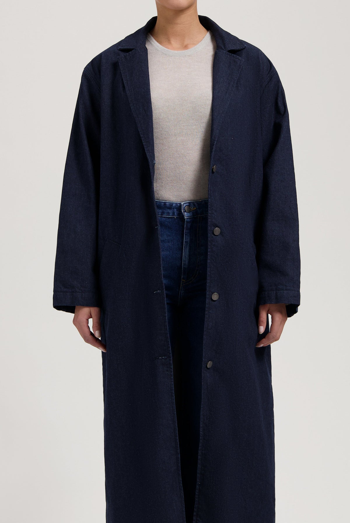 Long navy denim coat with button front, worn over a beige top and blue jeans, showcasing a minimalist casual style.