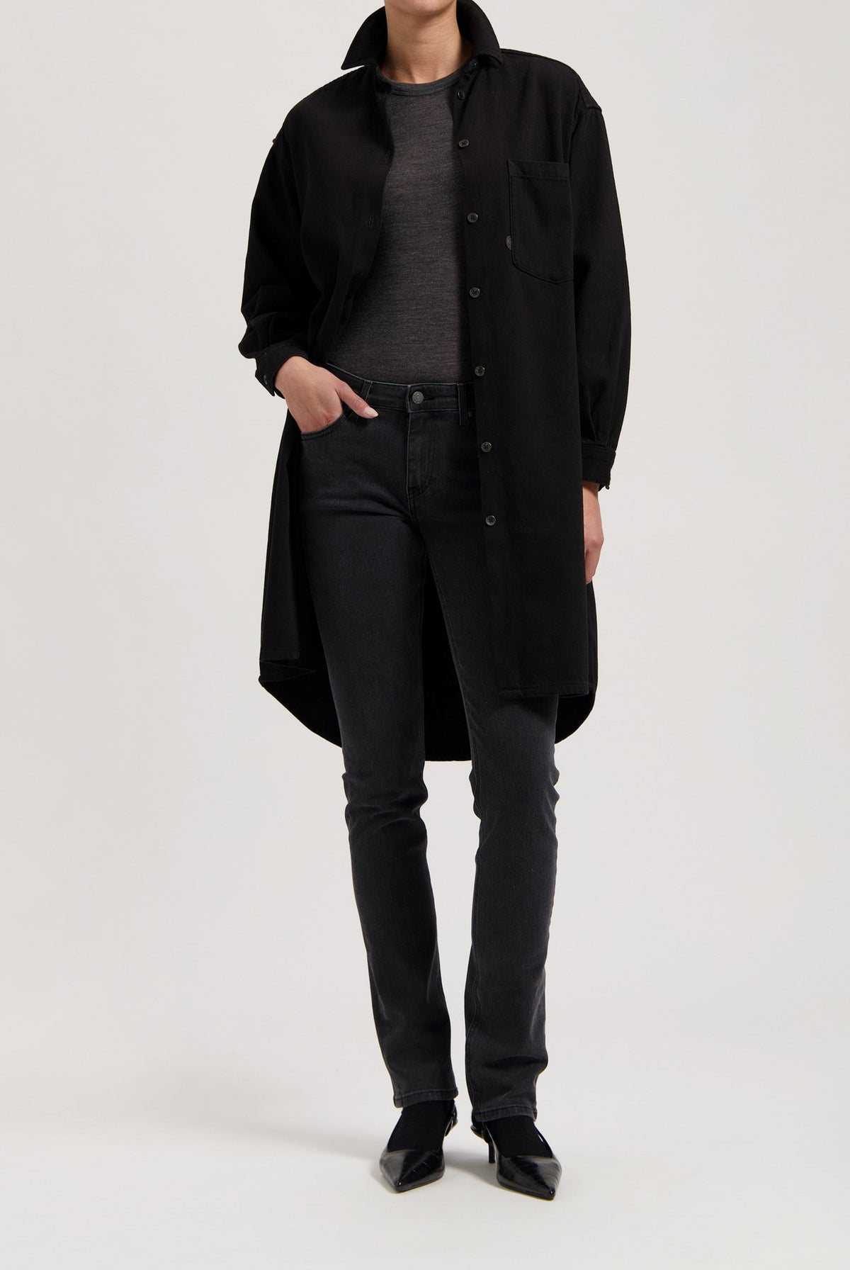 Model wearing a long black button-up coat over a gray top, paired with fitted black jeans and pointed black shoes.