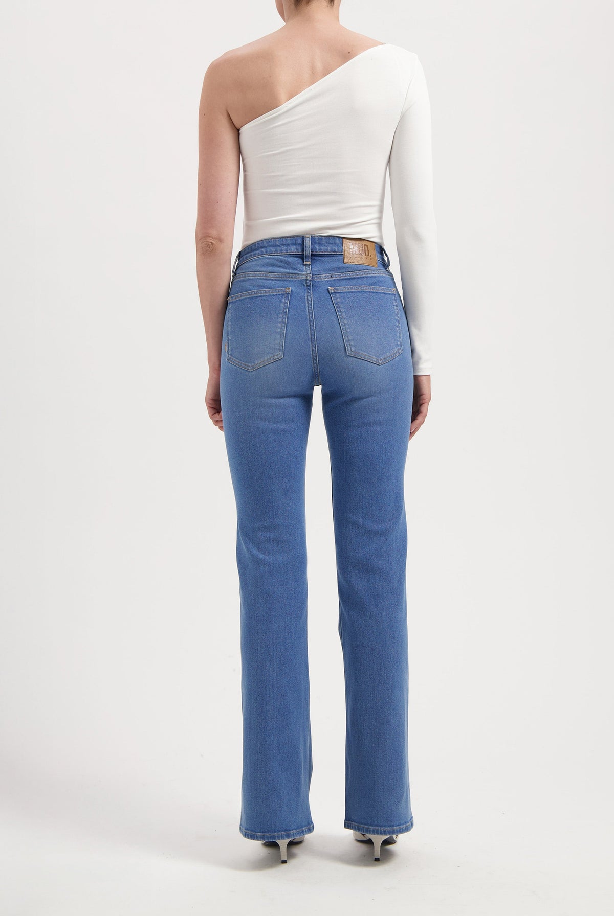 Model wearing blue high-waisted denim jeans with straight legs, paired with a one-shoulder white long-sleeve top.
